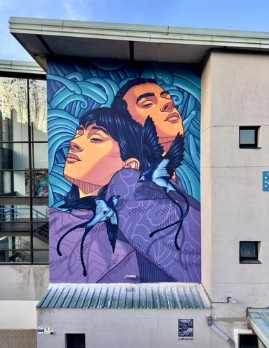 Streetartwall. A mural showing a young couple surrounded by blue grass and blue birds was sprayed/painted on the outside between the glass fronts of a modern two-storey school building. The entire mural is painted in shades of blue, purple and orange. The two figures have black hair and are wearing purple shirts. Their eyes are closed as they lean their heads against each other in the blue grass. Two dark blue birds with light blue chests and long tails flutter around in front of them.
The mural is finely drawn and rich in detail. Beautiful