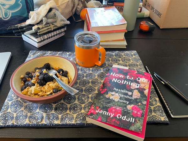 On a black wooden dining table, a brown-rimmed bowl of oatmeal with berries and nuts, an orange coffee mug with a lid, and a paperback book entitled "How to Do Nothing" by Jenny Odell, all sitting on a blue-and-white cloth placemat with circular designs. To the right is a black notebook and a black fountain pen. Behind it all the table has also some drawing/sketching pens, some other library books, a tape measure...