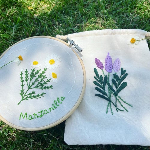 Flores de manzanilla bordadas con la palabra "Manzanilla" sobre un aro de madera y una bolsa de manta con unas lavandas bordadas sobre pasto.
