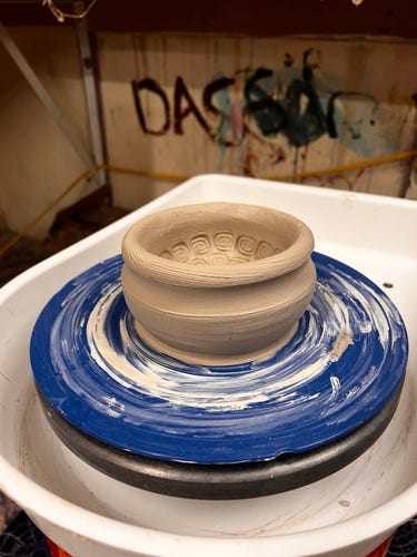 A freshly thrown clay pot on a spinning potter's wheel with decorative patterns inside, set against a backdrop of a paint-splattered studio wall.