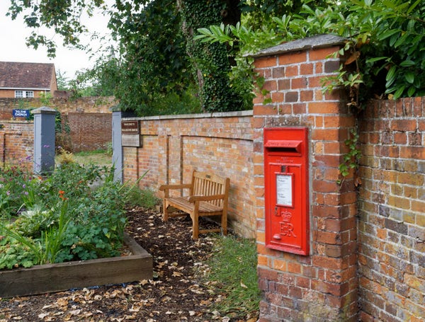 A red postbox, embedded at the end of a brick wall. There is a bench to its left, and some green leaves above the brick wall.