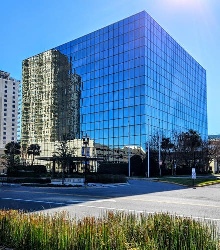 On a bright sunny day with clear blue skies,  sunshine bounces from nearby buildings onto a large square office building entirely covered in mirror-like windows, reflecting adjacent buildings and scenery.