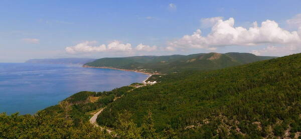 An aerial view of the world famous Cabot Trail in Nova Scotia.  The road winds along the Atlantic coast, surrounded by dense green forest.  The blue of the water is matched by the blue of the sky