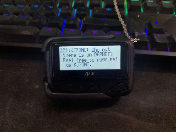 An alphanumeric pager standing up against a computer keyboard. Its screen reads "01:KJ7OMO: Who out there is on DAPNET? Feel free to page me! de KJ7OMO."