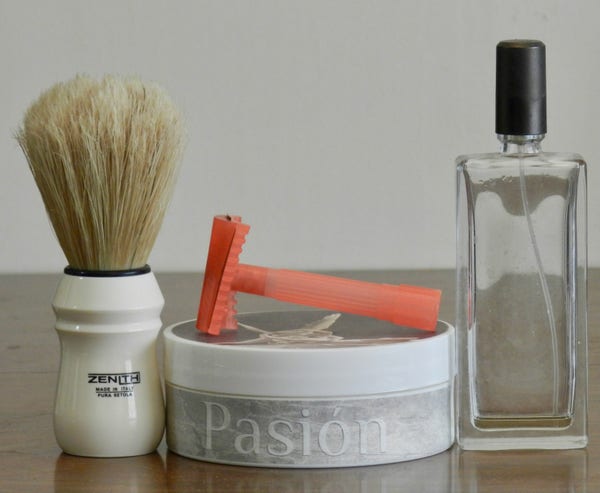A boar brush with a tan knot and white handle with "Zenith" printed in black stands next to a tub of shaving soap whose light-grey side-label has "Pasión" in white letters. Lying on top of the tub is a plastic slant razor in a faded crimson. Next is a rectangular clear-glass bottle with a tall black lid. The bottle has no label.