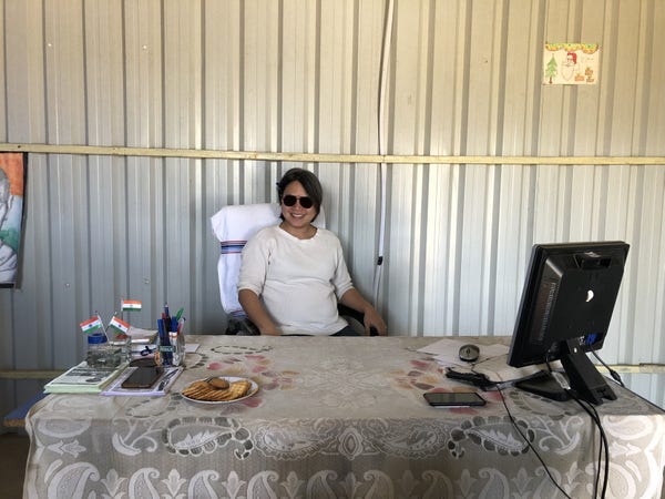 a photo of a east asian person wearing shades sitting at the principal's desk with indian flags at a rural school in india