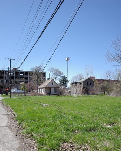 Color photograph looking over empty, grass-covered lots on the corner of a city block at a pair of disused houses on the next street, empty lots all around them, the end of a several-story high abandoned factory building looming up in the background behind them, a 5G tower standing up higher out of some open space behind the houses