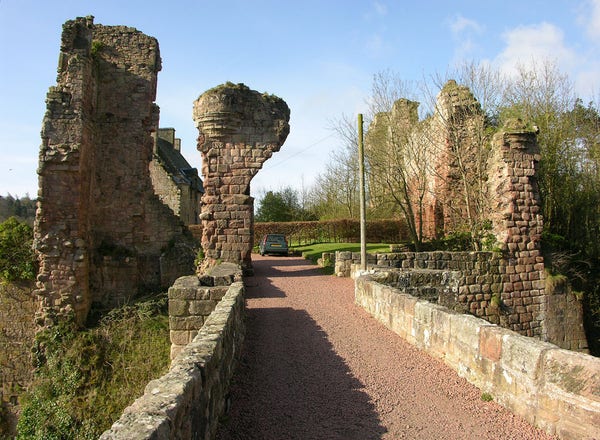 Rosslyn Castle. The image shows a gravel covered bridge with stone parapets leading into the frame. Beyond it is an area surrounded by fragments of castle ruins and, only partly visible on the left, a stone house. The ground drops away on both sides. The scene is in sunshine,