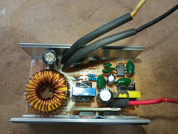 Color photo of a small electronic circuit board in an aluminum box. There's a yellow, black  and red wire coming out of it. Prominent on the board is a large round copper wire wound coil.