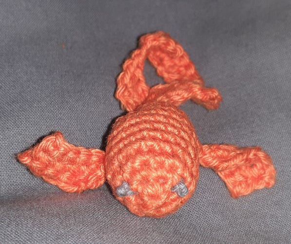 Ein selbstgehäkelter orangefarbener Fisch, dessen Anleitung KI-generiert ist, auf grauem Grund und graue Augen.
Die Schwanzflosse wirkt gewunden.
