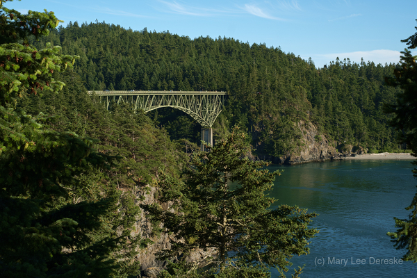 Deception Pass Bridge between Whidbey and Fidalgo Islands, Washington. Bridge is green arch steel, well over 100 feet tall. Hills are covered in pines, rock cliffs can also be seen, as well as a sand and driftwood beach.