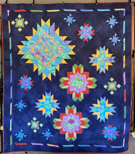 King size quilt with a dark blue background and various sizes and colors of star patterns and an orange binding.  Star colors include yellow, blue, pink, green, and orange.  You can just make out the meander quilt stitch on the background of the quilt.
