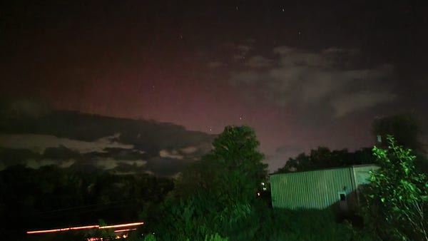High-gain long exposure shot of the sky at night featuring some dark clouds silhouetted against a green and pink glow