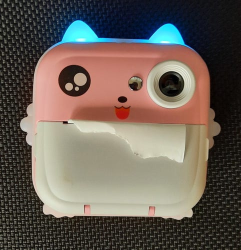A pink and white toy camera formed like a cat, with one cat eye used for the camera, and with blue glowing silicon ears. The bottom half of the face is a door holding a roll of thermal paper.
