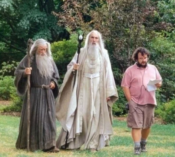 Two actors as wizards in Lord of the Rings - in full costume with beards and staffs - listening while walking to director Peter Jackson casually wearing a polo shirt and shorts.