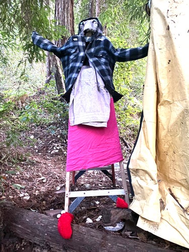 A towering, eerie figure with a ladder for bones, bright red booties, a pink skirt, an apron made of a pale dirty towel, a big flannel overjacket with hood and arms outstretched and glow-in-the-dark bug stickers at the shoulders, and a face made from a white-tiger pillowcase with chin propped up by a rock