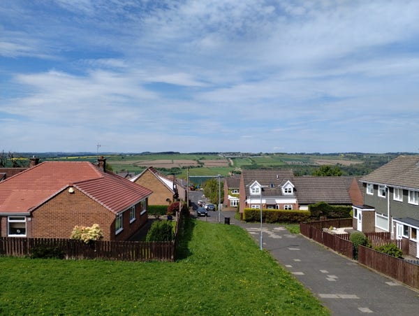 A view over a small square of houses. In the middle is a paved footpath and a small grassy bank, covered with yellow dandelions as it's May. In the distance, the view is over a rural valley, farmland on a mixture of greens, yellows and browns. The sky is bright with a patchwork of blues and light high clouds.
