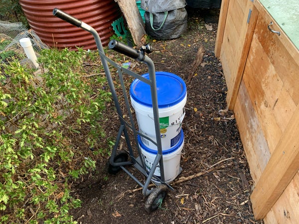 A basic steel sack-barrow style hand cart carry two large catering buckets, parked near a wooden compost bay