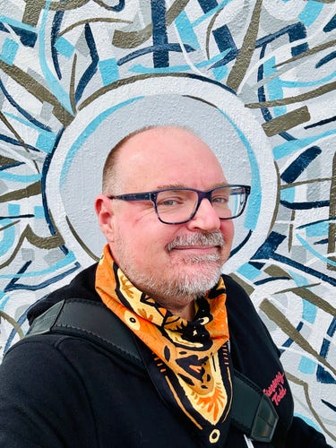 A selfie of me wearing a Sweeney Todd hoodie and a tan & yellow neckerchief. I’m standing in front of a very colorful mural. A gray, blue, white and brown circle forms a painted halo directly behind my end.