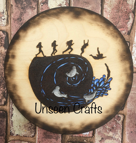 Circular wooden plaque with burned design showing stages of running and diving into the water. Three grey seals in a spiral. Elements in the water are painted in blue and white. The edge of the plaque has been scorched all the way around.
