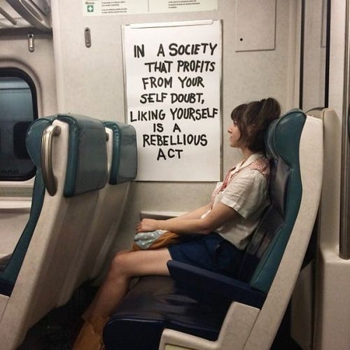 Woman seated on public transit. On the wall next TO her is a frame for holding an advertising poster. The ad has been replaced with a white handwritten sign that reads “IN A SOCIETY THAT PROFITS FROM YOUR SELF DOUBT, LIKING YOURSELF IS A REBELLIOUS ACT”