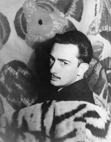 Image en noir et blanc représentant Salvador Dali de dos. Il porte un manteau à motif zébré. Le fond présente un grand motif floral distinctif. Ses cheveux sont soigneusement peignés et coiffés.