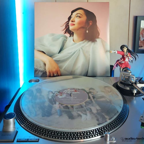 A clear vinyl record sits on a turntable. Behind the turntable, a vinyl album outer sleeve is displayed. The front cover shows mxmtoon looking off to the left. Her arm is laid across a pillow. 