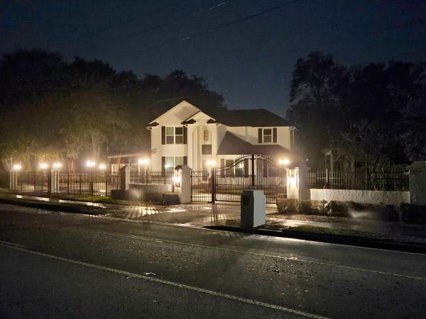 Late night view across a wet roadway in the dark at a freshly refurbished hundred-year-old farm house beyond a brick wall and black wire fence, illuminated with numerous torch like lamps surrounding the property.