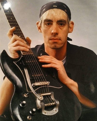 Greg Sage of the band Wipers, a blonde haired guy with a black head scarf in a black shirt holding a black guitar.
