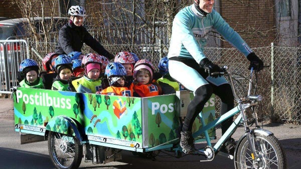A bike with a “truck bed” with a lot of small seats for children. Filled with kids with bike helmetsz