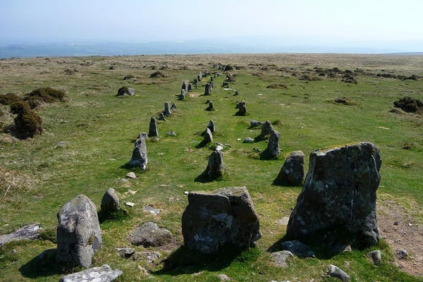 Three parallel rows of granite standing stones head away from the viewer, curving slightly to the right as they run down a gently sloping hill. Three taller stones mark the ends of the rows nearest the viewpoint. None of the stones are very big, but the rows are well preserved with over a dozen stones visible in each. The terrain is short grass. Faint blue hills form the distant skyline backdrop. The sky is clear blue and it's very warm.