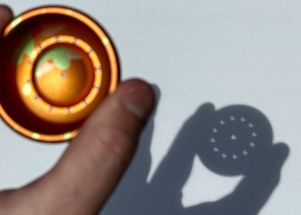 A photo of half a yo-yo being held near a piece of paper on a sunny day. The yo-yo has little holes drilled in it, and when the light passes through those holes you can see the shape of the moon as it passes in front of the sun. 