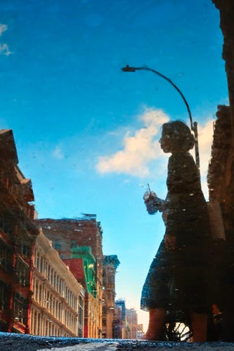 A scene reflected in a puddle: a silhouette of a person is reflected walking from one side of the street to the other.

On the left side of the image is a line of buildings shrinking into the distance. The person is on the right side. Behind the person is a street light and tall buildings.

The person is holding a plastic cup with a dome lid and a straw. They have long hair kept in a bun at the back of their head.

At the bottom of the image is the asphalt edge of the puddle reflecting the scene above.