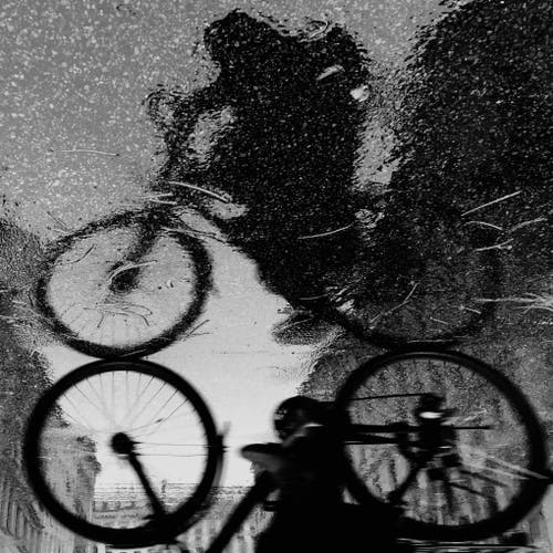Schwarz-Weiß-Bild, das die Reflexion eines Radfahrers und von Fahrradrädern auf einer nassen Oberfläche zeigt, wodurch ein abstrakter und künstlerischer Effekt entsteht.