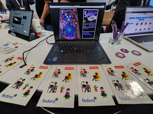 Fedora and CentOS booth at Red Hat Summit with a Lenovo display, ThinkPad X1 Carbon, and Slimbook Fedora 1 from left to right; with a smattering of stickers and swag available