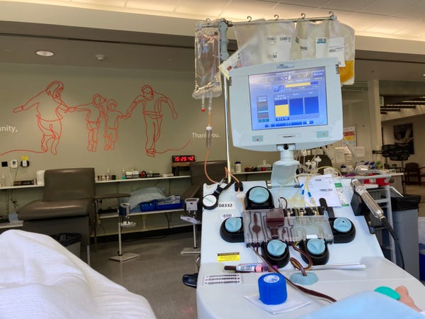 Blood donation apheresis machine with lots of tubing. There is a mural on the wall in the background made of curving red lines showing two adults holding hands with two children. 