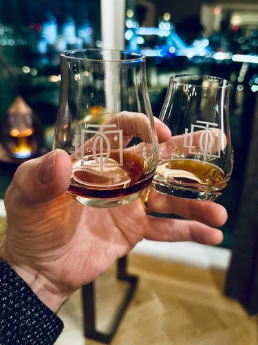 A hand holds two small glasses of liquid. One is a rich, deep chestnut brown and the other a golden light amber. Guess which one is which?