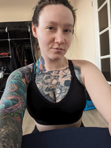 selfie of a person with white skin mostly covered in tattoos, crouching down next to a bed with messy pulled back hair, a black v neck bra, and black pants 