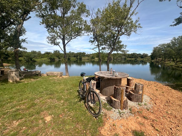 un vélo contre une table de picnic, nourriture et boisson poussée dessus, devant un lac bordé d'arbres suis un ciel bleu radieux