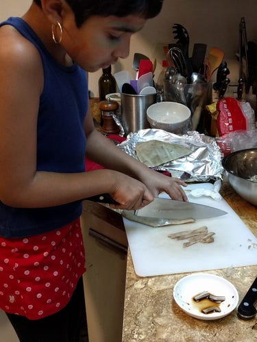 Kid standing on a stool, and cutting slices of something with a chef's knife on a white cutting board.