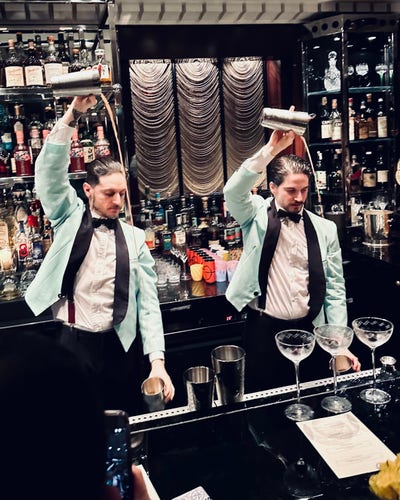 Two bartenders busy rolling drinks - that is, pouring the contents of their cocktail shakers from one half, held high above their shoulders, into the other half which is held down by their hips. They are doing this in unison, in a snazzy looking hotel bar. They are wearing little duck-egg blue jackets with black satin lapels over white shirts with black braces (suspenders for you Yanks) and black bow ties. 
