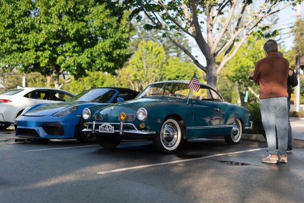Dark teal classic coupe, next to a blue sports coupe. Spectators stand around the car.