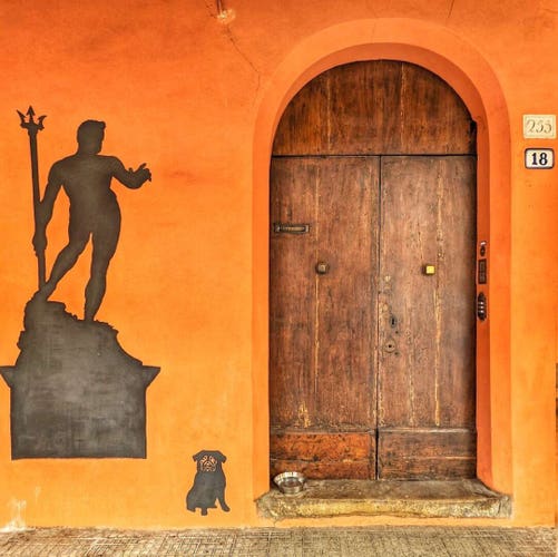 Worn wooden door with two panels each with a square metal door knob. There is a "letter" slot near the top of the left panel. The facade is bright orange. There is a statue of Neptune painted in black paint to the left of the door that resembles Neptune on Piazza Maggiore in Bologna. There is a small pug painted in black sitting next to the door. There is a metal doggie dish filled with water sitting in the