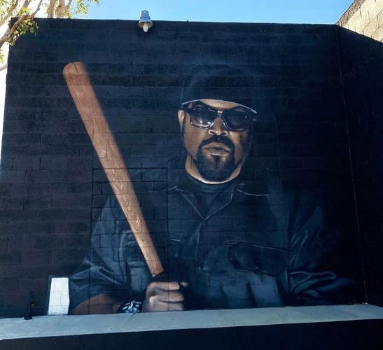 Streetartwall. The dark portrait of the black rapper "Ice Cube" has been sprayed onto a black exterior wall. The man has a full beard, wears sunglasses, a black baseball cap and is dressed in black.  He is standing there with a baseball bat in his hand. A little light falls on his face. He looks menacing. 
Info: The singer was present at the spray session and killed a computer for it.