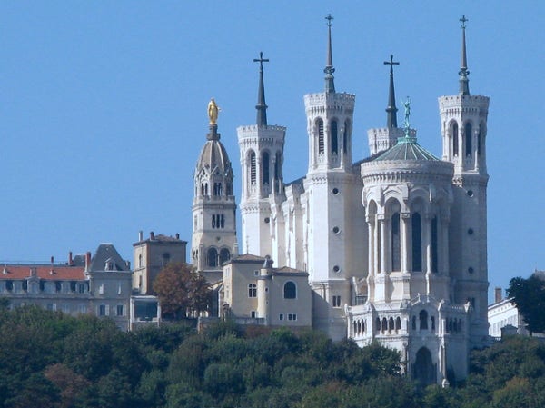 a photo of a large church its walls are eggshell white, it has large copper green spires on the top with crosses, on one of the domes is a golden statue, the sky is blue, there are trees and buildings in the foreground and background 