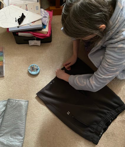 A person sewing a dark piece of fabric, with pins in a pin cushion and sewing materials scattered around on the floor.
