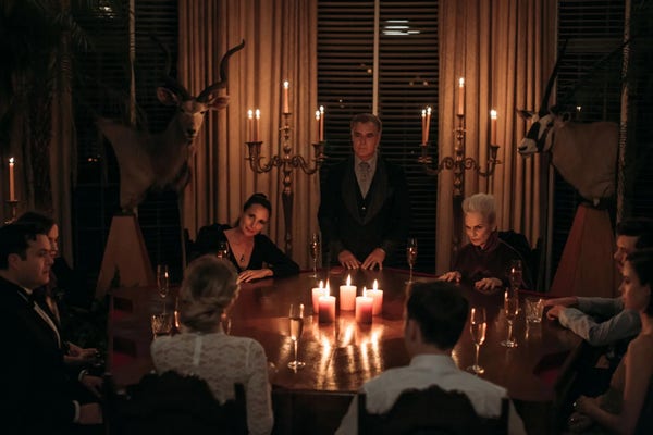 Still from the horror movie Ready or Not where a family of heirs to a boardgame company fortune who hunt and kill anyone that marries in to the family are gathered around a table looking at a new bride