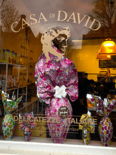 Store front of an Italian deli in Amsterdam. The bottom of the window frame is filled with large Easter themed egg like pacakges. One in the middle is extra large and it's wrapping paper goes up to meet the logo of the deli and is the head of a classical figure.