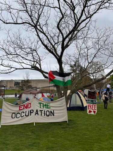 Tents being constructed on the university lawn.  A huge windbreaker reading END THE OCCUPATION in the foreground of the image.