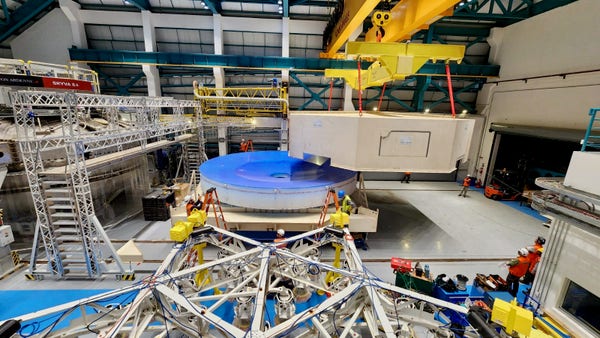 A thick, 8.4-meter wide white disk of glass with a blue protective coating on the top surface is sitting on a white platform. A white box top that covered the glass is being lifted away.